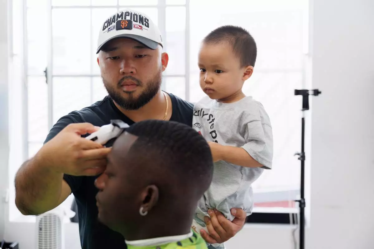 Jack Chang cuts Isaac Obamehinti's hair while holding his 2-year-old son Cairo Miu at his studio on Monday, July 13, 2026, in San Francisco. (AP Photo/Juliana Yamada)