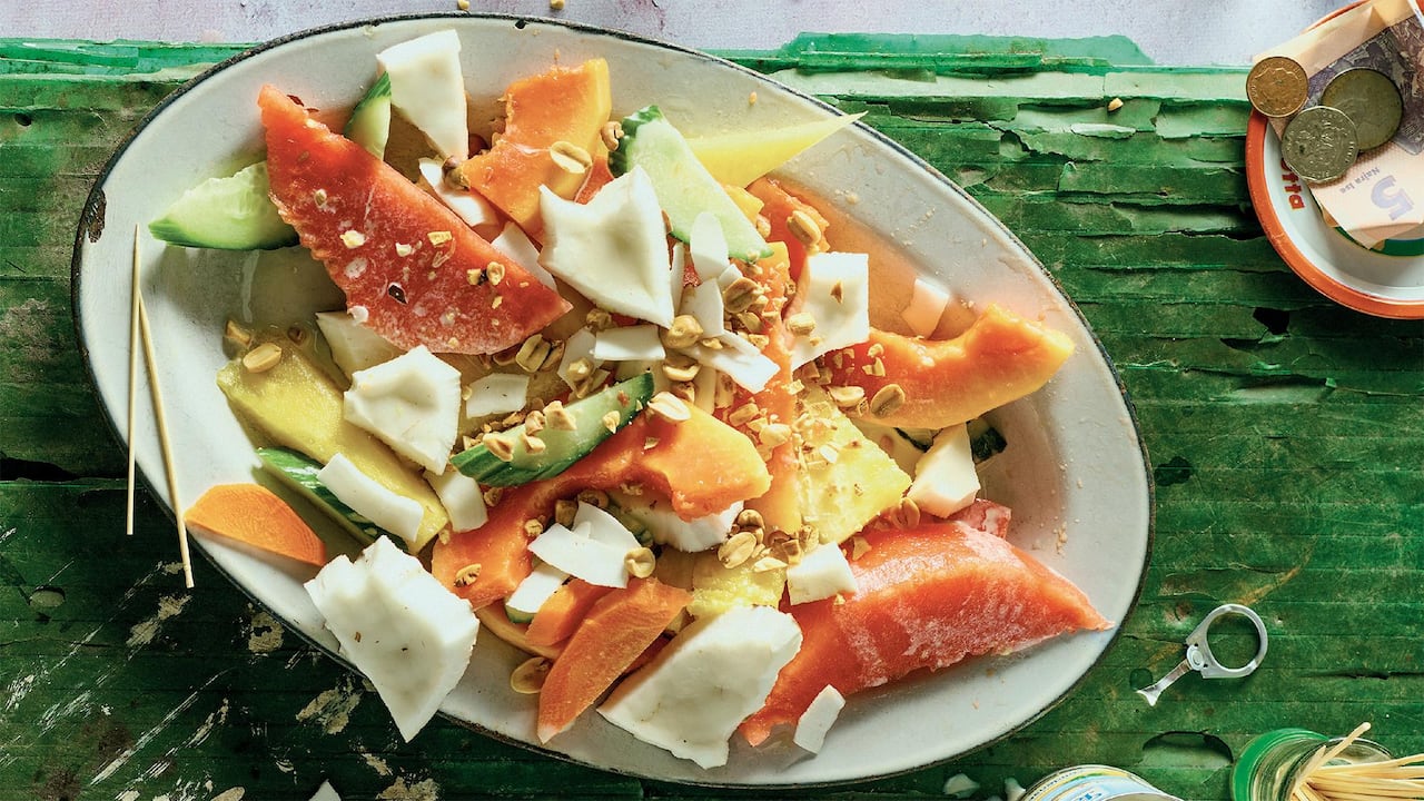 Top down shot of a white platter of fruit salad with large chunks of pineapple, papaya, cucumber, coconut and carrots and topped with peanuts. 