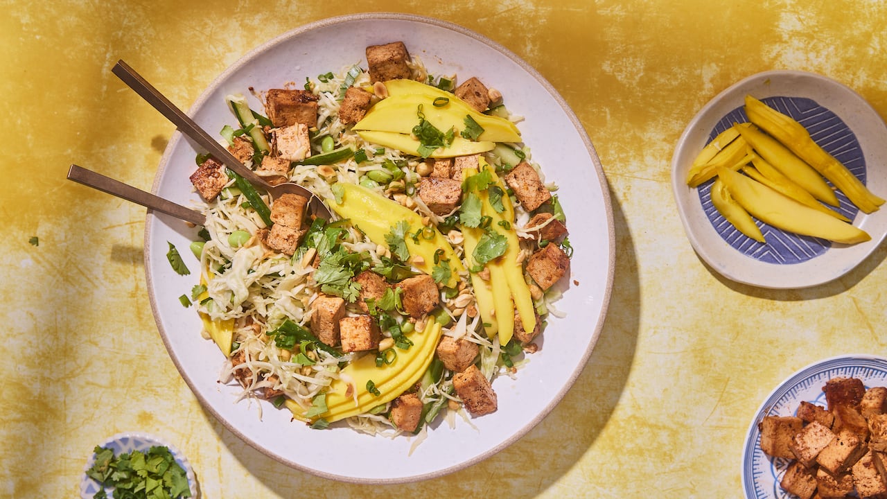 Top down shot of a big bowl of mango salad with shredded cabbage, edamame and cubes of tofu. It sits on a yellow surface with small bowls of ingredients beside it.