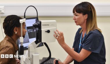 A nurse giving a woman an eye screening test. Both are seen side on. The nurse, on the right, has brown hair, and is wearing a blue uniform. One hand is adjusting a dial on a machine, similar to a large camera. The woman to the left is looking into the machine with her chin resting on a frame. She has dark braided hair and is wearing a brown leather jacked. A screen can be seen to the side of but the images it is displaying cannot be made out.