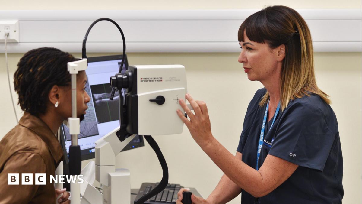 A nurse giving a woman an eye screening test. Both are seen side on. The nurse, on the right, has brown hair, and is wearing a blue uniform. One hand is adjusting a dial on a machine, similar to a large camera. The woman to the left is looking into the machine with her chin resting on a frame. She has dark braided hair and is wearing a brown leather jacked. A screen can be seen to the side of but the images it is displaying cannot be made out.