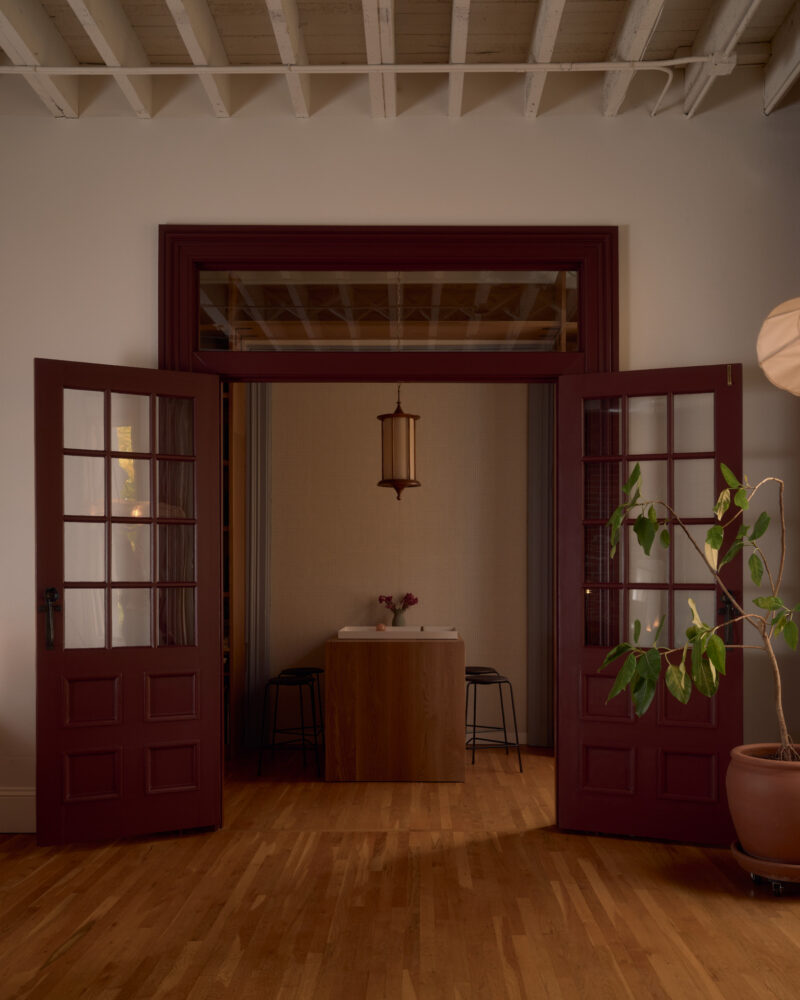 Open double doors reveal a small table with two stools in a softly lit room; a potted plant stands to the right on a wooden floor under an exposed beam ceiling.