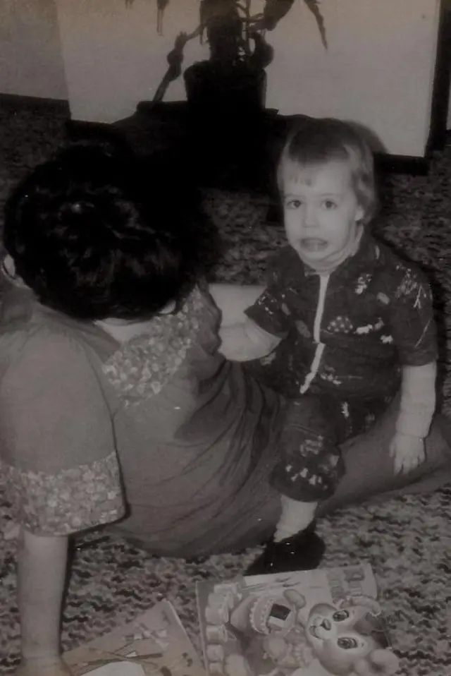In a black and white photo, a small child (Nina) sidon for her mama lap on di floor. Di mama dey look down so we no see her face. Two children books lie on di floor in front of dem.