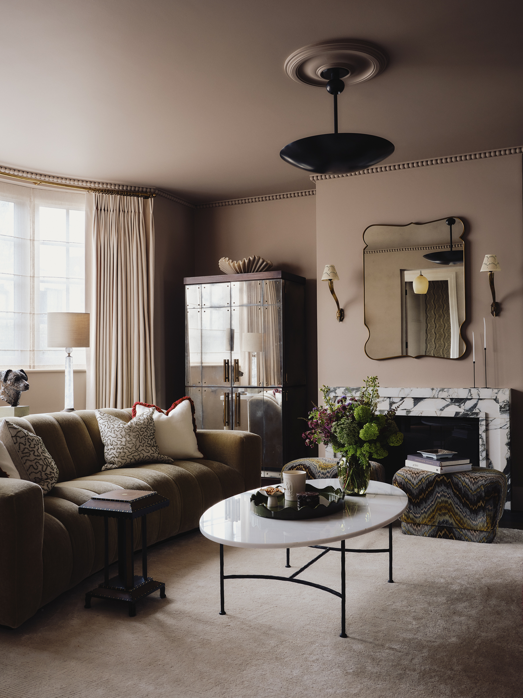 cozy living room with taupe color-drenched walls, white curtains, marble fireplace with mirror hanging above, mirrored cabinet, tan sofa, white marble coffee table with black legs and beige rug