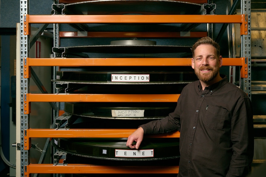 Jeremy Fee standing next to reels of film with Christopher Nolan titles on them including Inception and Tenet
