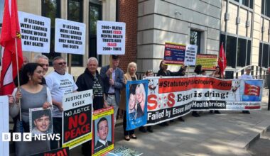 Protesters stand outside Arundel House in Central London. They are holding a big horizontal banner that says "SOS Essex is dying!" Some are also holding posters of their relatives that have died. They are standing outside the front of the mock-Tudor building. There is a row of about 30 people.