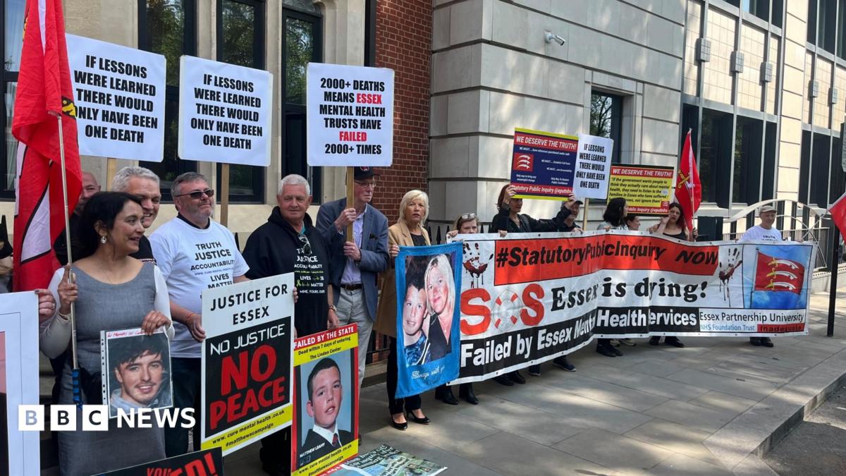 Protesters stand outside Arundel House in Central London. They are holding a big horizontal banner that says "SOS Essex is dying!" Some are also holding posters of their relatives that have died. They are standing outside the front of the mock-Tudor building. There is a row of about 30 people.