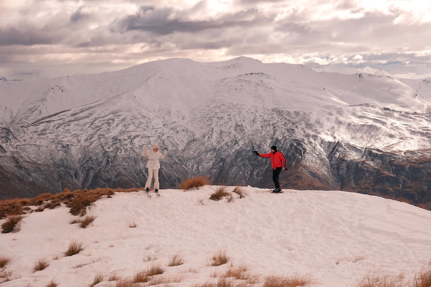 Wide shot of snow-covered mountains, Paul Williams with his hands in the air, a man in a red snowsuit pointing a gun at him