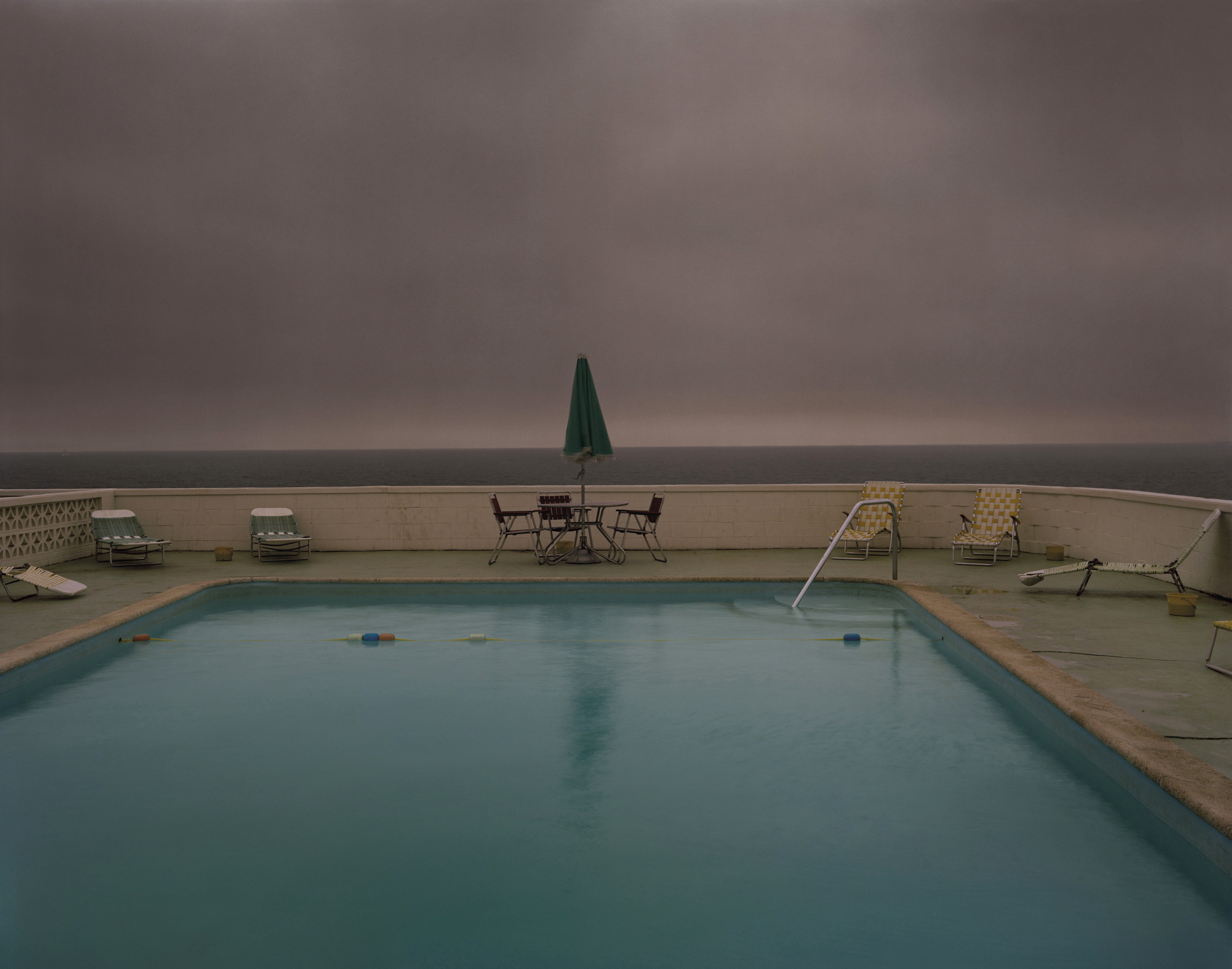 A rectangular motel swimming pool sits empty under a heavy grey storm sky, with vacant lawn chairs and a folded green umbrella lining the poolside terrace.
