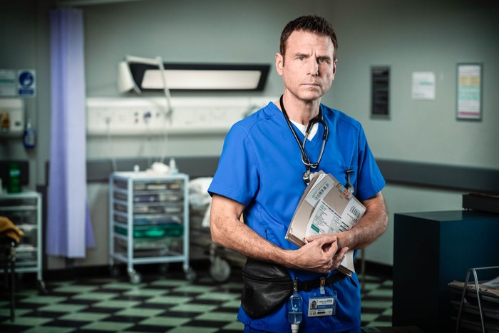 336712,TITLE:Casualty - Portraits 2021 Jason Durr as David Hide in blue scrubs holding a folder in Casualty in 2021.