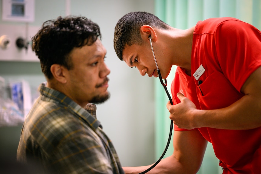 A TV still of Ioane Sa'ula, mid-20s, Samoan Australian, in orange scrubs, holding a stethoscope to Aliki Matangi's chest.