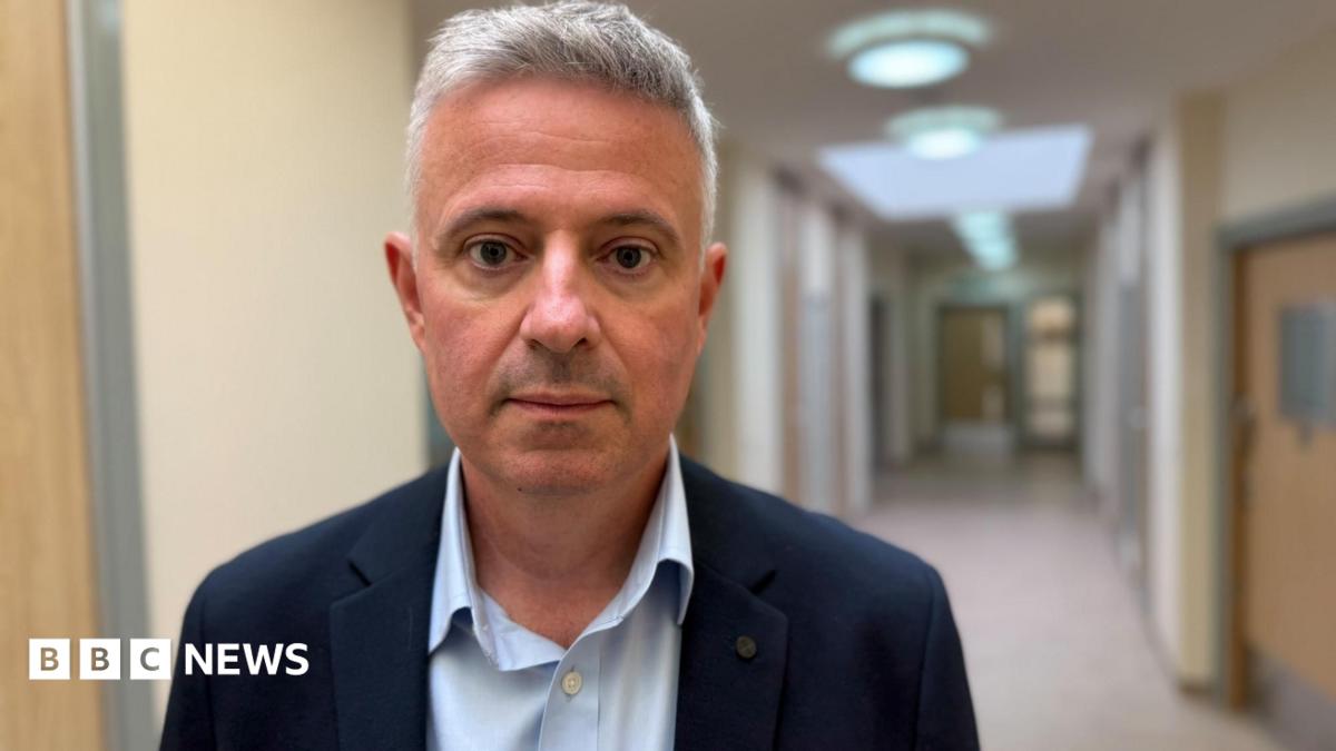 Paul Scott looks at the camera. He is standing in front of a corridor near the trust's headquarters. It is an inpatient unit. You can see a new beige corridor with light colured wood doors. Scott is wearing a navy suit jacket with a pale blue shirt