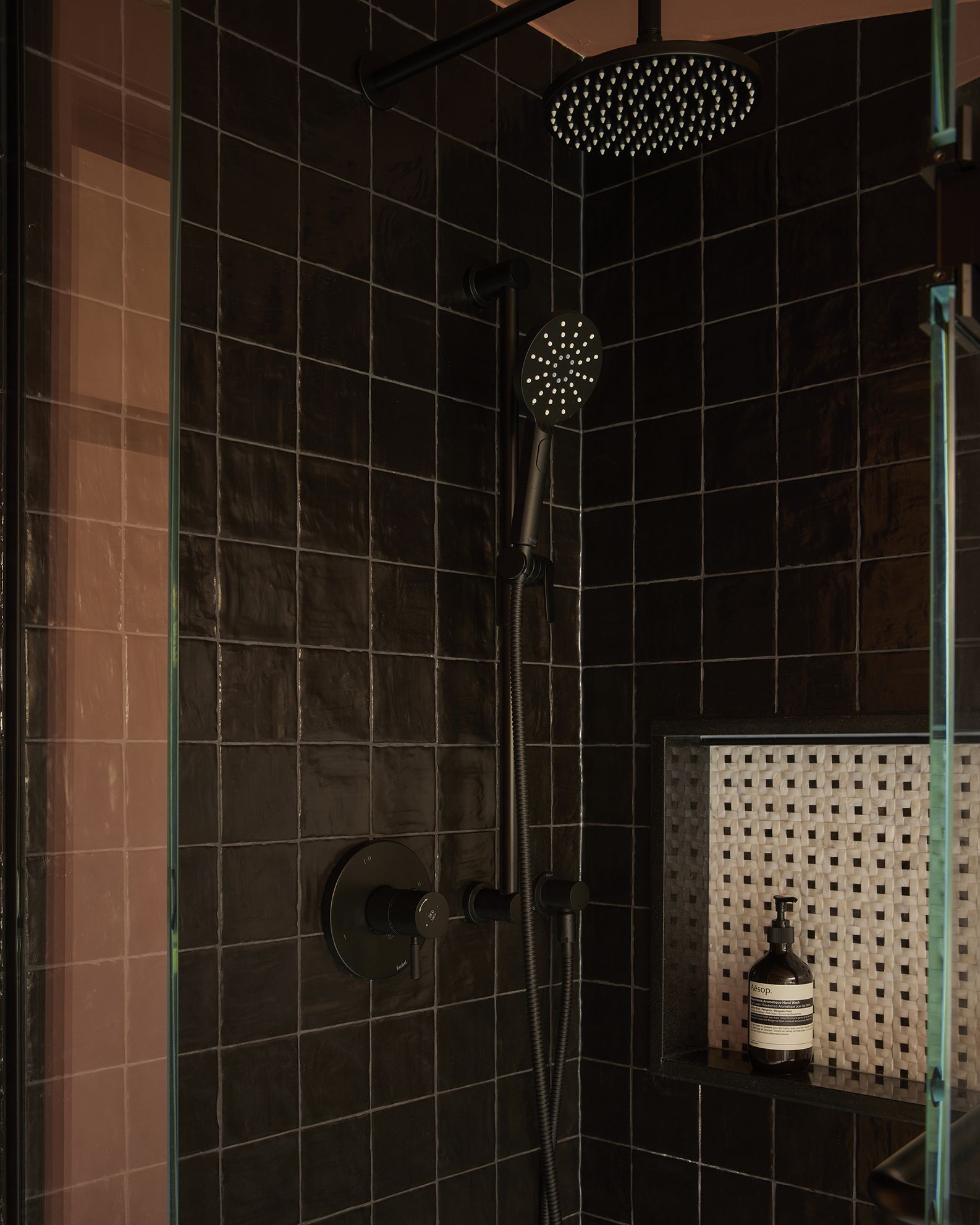 A brown tiled shower with Aesop products, black shower head and over-head shower, and see-through glass.