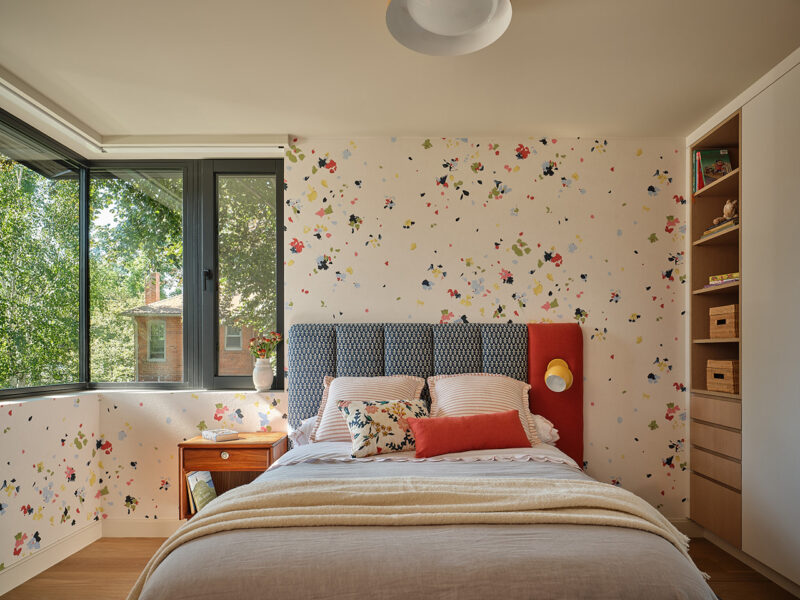 A cozy bedroom with floral wallpaper, a bed with colorful pillows, a wooden nightstand, built-in shelves, and large windows overlooking green trees embodies the thoughtful design seen in a Gillian Gillies Net Zero Ready Home.