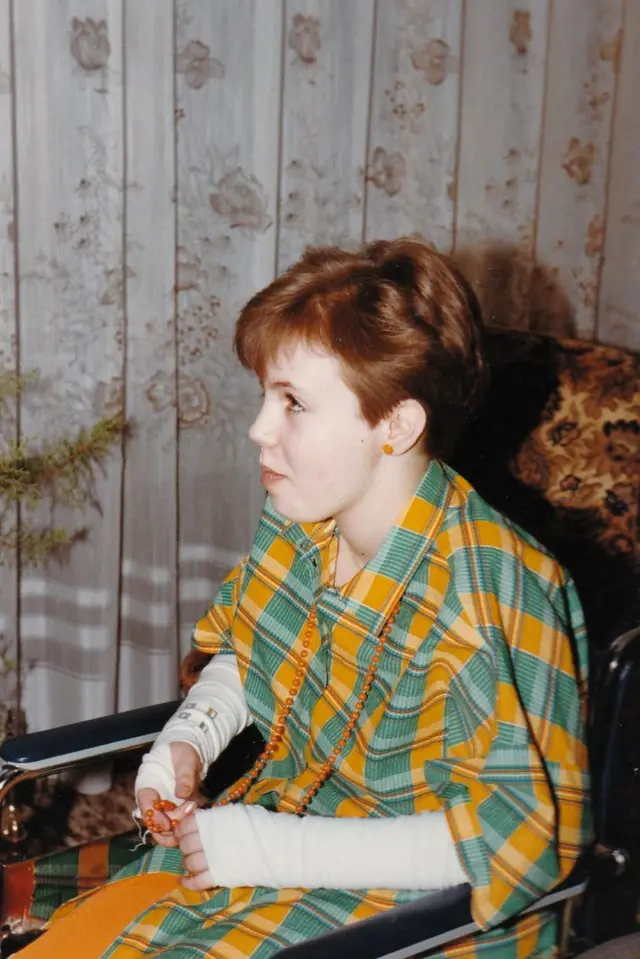 A colour 1982 photograph show 11-year-old Nina sidon for wheelchair in a living room. Di pesin dey wear a matching green-and-yellow checked outfit over a light-coloured long-sleeved top and dey hold a string of orange beads. Lace curtains cova one window for di background, and a patterned armchair dey partially visible beside di wheelchair.