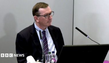 Brian O'Donnell sits behind a desk at the Lampard Inquiry at Arundel House in London. He is wearing a navy and burgundy tie and a black suit jacket, and a black laptop sits in front of him at the table. There are two bottles of water on the table and a microphone in front of him with a box of tissues.