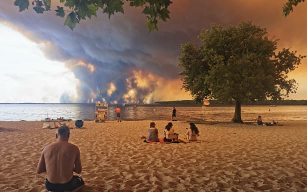 Beachgoers sit on the beach of Moutchic in Lacanau, southwestern France on 24 July, 2026, as clouds of smoke rise into the sky from a wildfire raging in Gironde, southwest France.