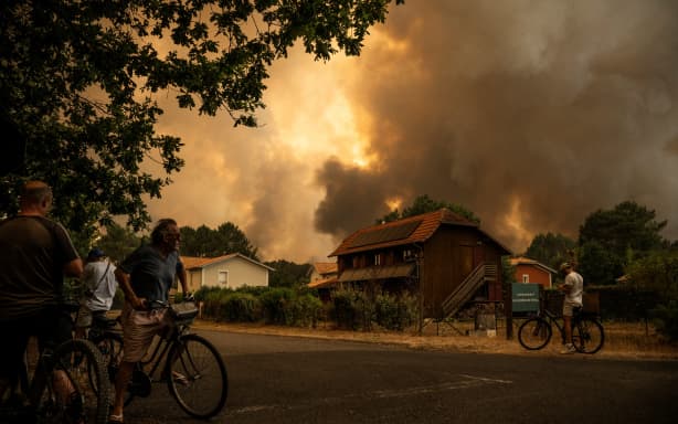 Residents on their bicycle look at smoke rising behind a residential area during a wildfire near Lege-Cap-Ferret, southwestern France on July 23, 2026. The fire raging in the northern part of the Arcachon Basin in Gironde, southwest France, has swept through more than 3,400 hectares, according to the latest figures released by the prefecture. More than 20,000 residents and tourists have been evacuated from this part already devastated by flames in 2022. France has been battered by three heatwaves since May as part of increasingly common extreme weather episodes linked to climate change, causing excess deaths, drying up rivers and fuelling wildfires. Scientists have long warned that climate change caused by human use of fossil fuels will make periods of drought more intense and longer-lasting, creating the ideal conditions for wildfires. (Photo by JULIEN DE ROSA / AFP)