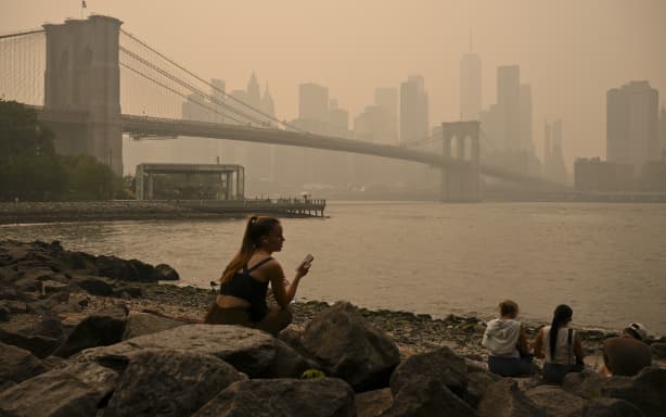 People sit near the Brooklyn Bridge as wildfire smoke from Canada causes hazy conditions on July 16, 2026 in New York. Out-of-control wildfires were raging Thursday in the Canadian province of Ontario, prompting evacuations and sending dangerous smoke billowing into the United States where millions of people were exposed to the unhealthy air. US states near the Canadian border including Minnesota, Wisconsin, Michigan and Illinois were particularly choked, as forecasters predicted the Northeast including New York would also see air quality continue to deteriorate. (Photo by ANGELA WEISS / AFP)