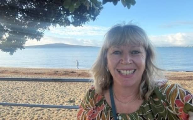 Sarah Bloxham stands on a boardwalk in Auckland with a beach and Rangitoto in the background.