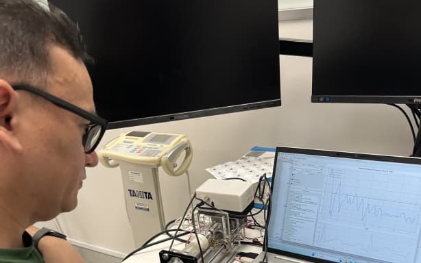 Dr Tet Chuan Lee sitting in front of a computer screen showing blood pressure oscillation lines. Hovering over the mousepad is his right hand and he has a prototype blood pressure cuff on on his index finger.