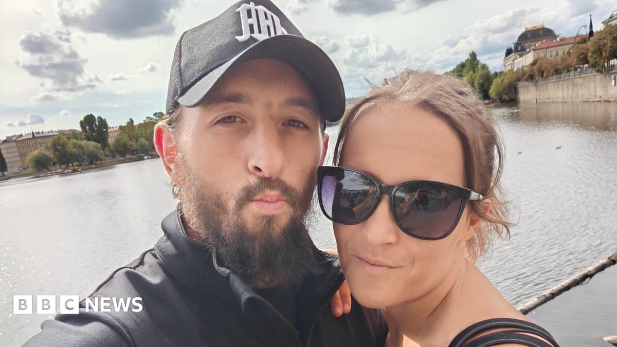 A selfie of a man and woman. On the left in the foreground, Kelvin Farmer has a brown beard and is wearing a dark coloured cap. On the right is Kayleigh, wearing dark rimmed sunglasses and her brown hair is tied back. In the background is a large body of water which could be a river or lake, and a cloudy but sunny daytime sky.