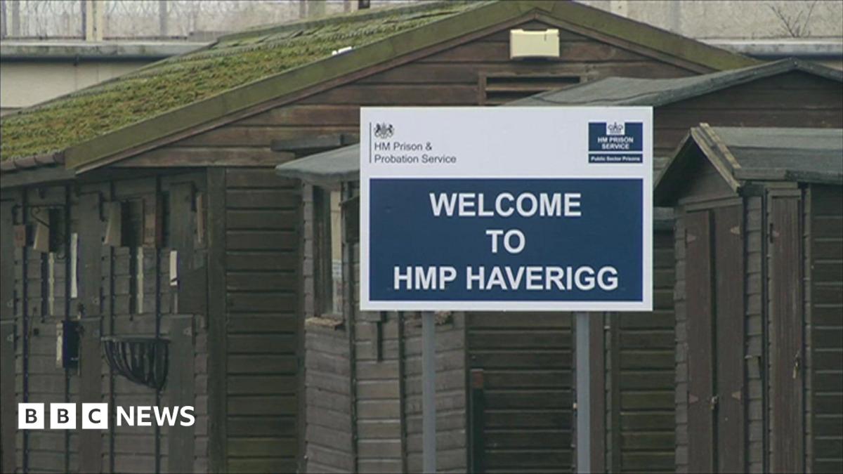 A blue-and-white sign saying "Welcome to HMP Haverigg". It is in front of hut-style buildings painted brown outside the main gate of the prison.