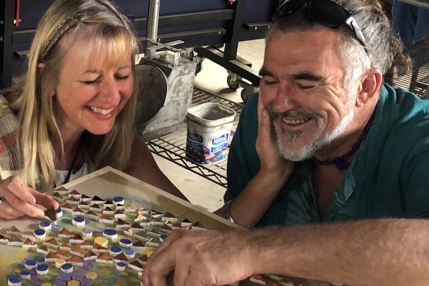 A man and woman smile as they work on a mosaic of colourful tiles.