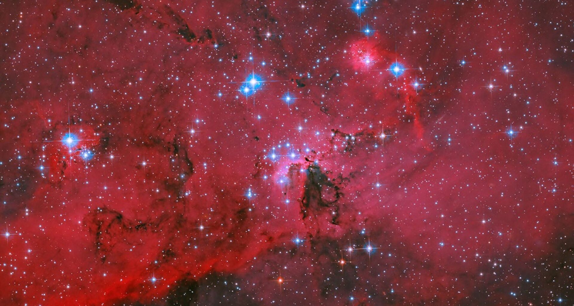 A Hubble image of blue and white stars are sprinkled throughout a landscape of red clouds of gas and dark, swirling lanes of dust. Many additional stars are visible in the dark background of space.