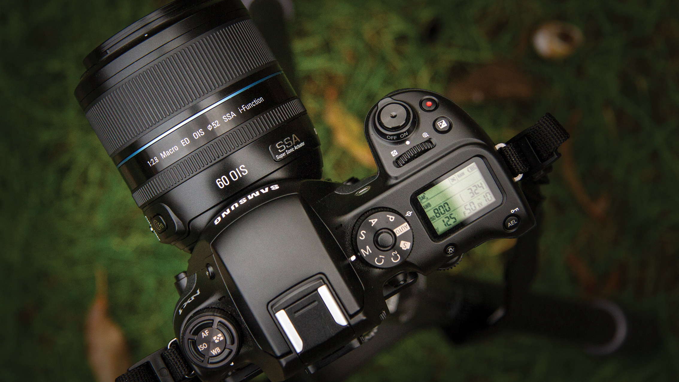 Black Samsung DSLR camera with 60mm macro lens mounted, showing top controls and display, set against a blurred green grassy background