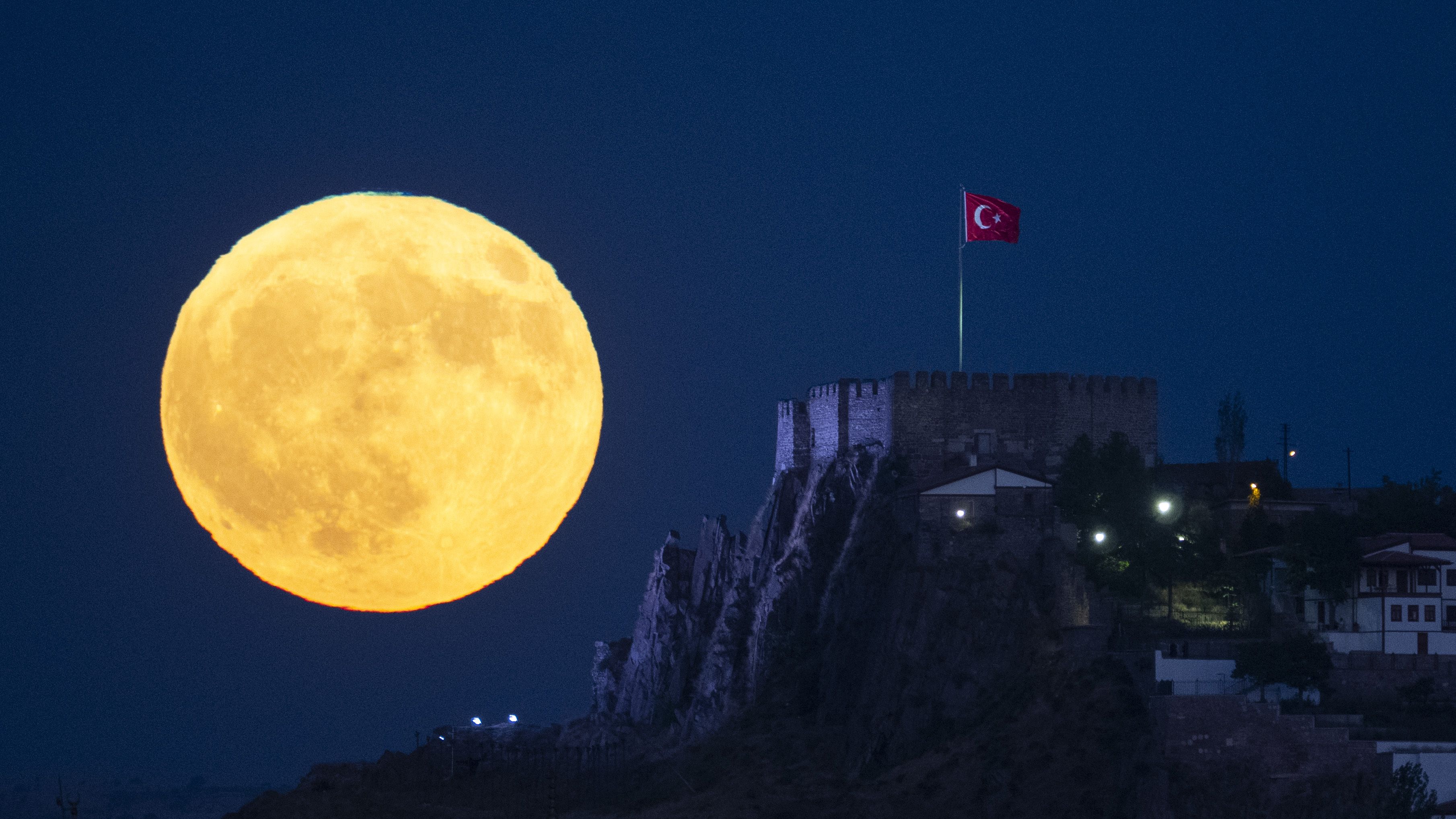 An orange full moon is photographed glowing to the left of a castle on a rocks hillside in a dark blue sky.