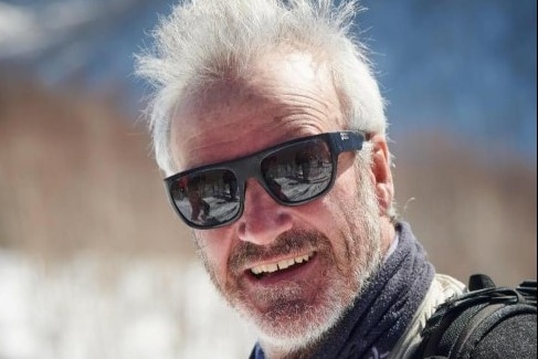 Grey haired man standing in the snow and wearing glasses smiles at the camera.