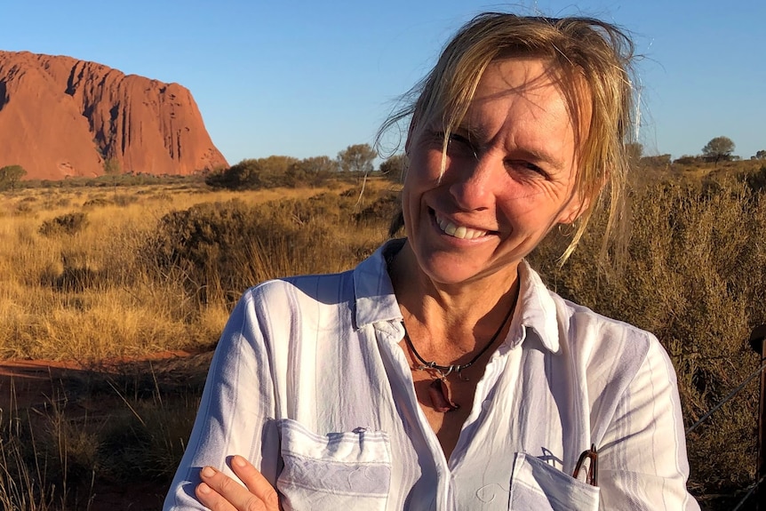 Sarah Legge smiles, stands in front of Uluru, has windblown hair.