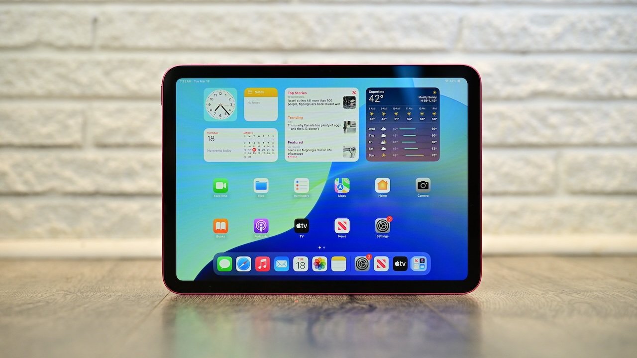 Apple iPad tablet with colorful home screen and app icons, standing upright on a wooden surface, with a white brick wall in the background