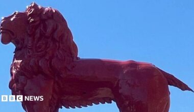 A wooden red lion sculpture stands atop a building, its mane and muscular form gleaming in the sunlight against a clear blue sky. Its tail has broken off.