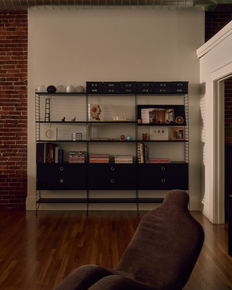 Black metal bookshelf with drawers, books, decorative items, and boxes stands against a white wall with exposed brick and wooden floor. A brown chair is partially visible in the foreground.