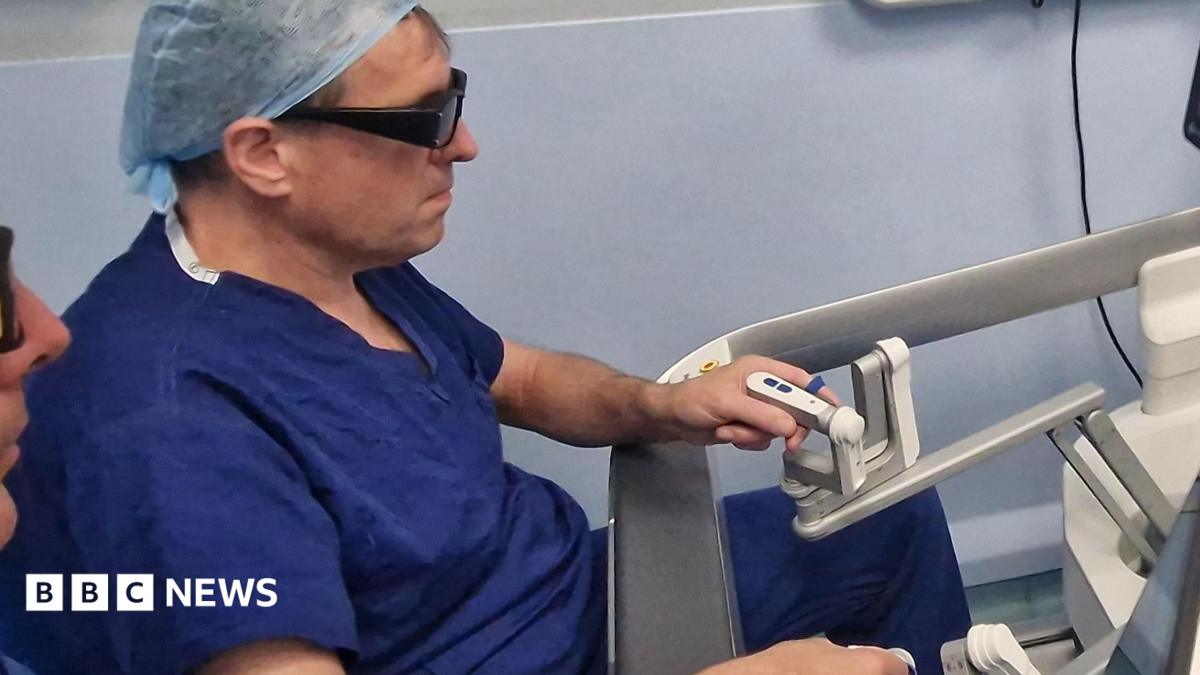 A medic wearing blue surgical scrubs and a head covering is sitting at a screen which shows an operation. He is holding two controls to direct the operation.