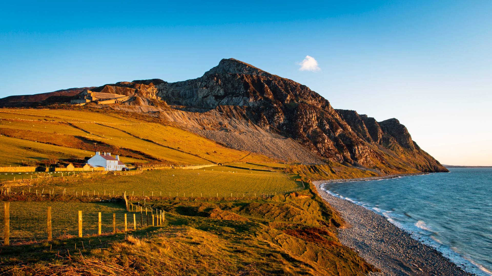 A coastal landscape at sunset features rolling green fields, a white cottage, rocky cliffs, and a pebble beach bordering calm blue sea under a clear sky.