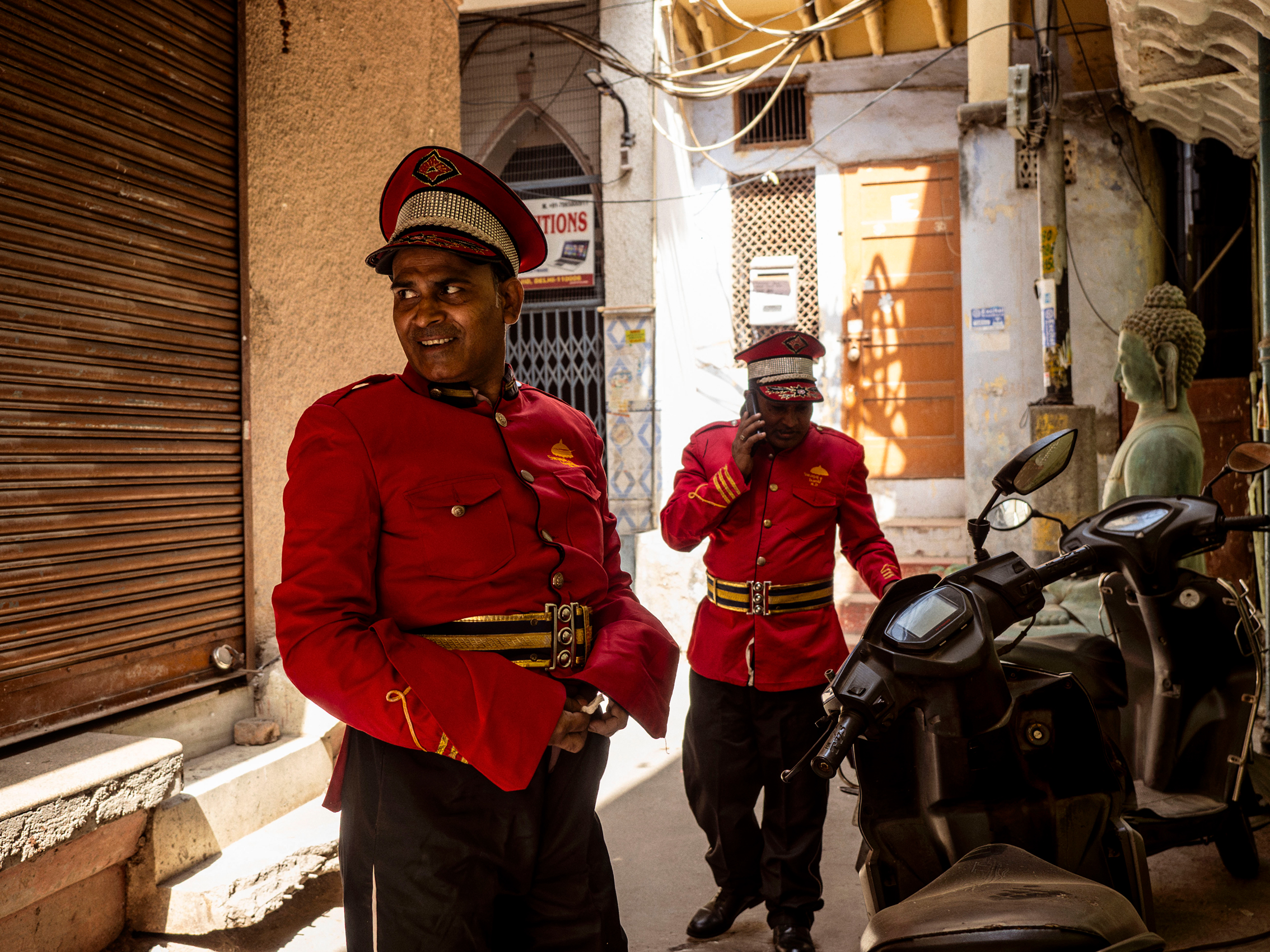 Two men in red uniforms and hats are standing in a side street in India with scooters next to them, one man is holding a smartphone, the other places his hands on his stomach