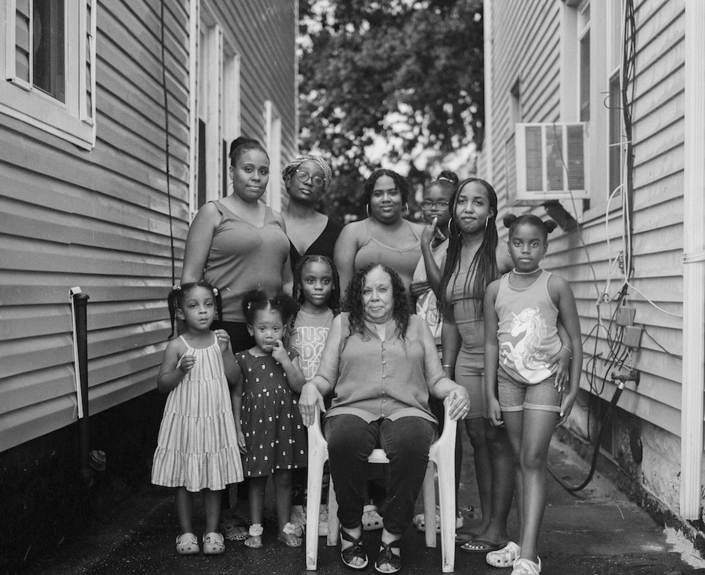 A black-and-white group portrait depicts a multigenerational gathering of girls and women posing closely together outdoors in a narrow alleyway between two buildings