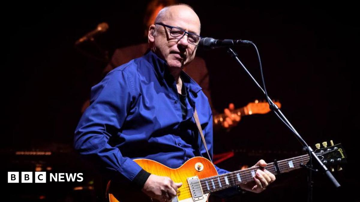 Mark Knopfler performing onstage. He is a bald man with glasses, wearing a blue shirt, and playing a guitar. A microphone is in front of him.
