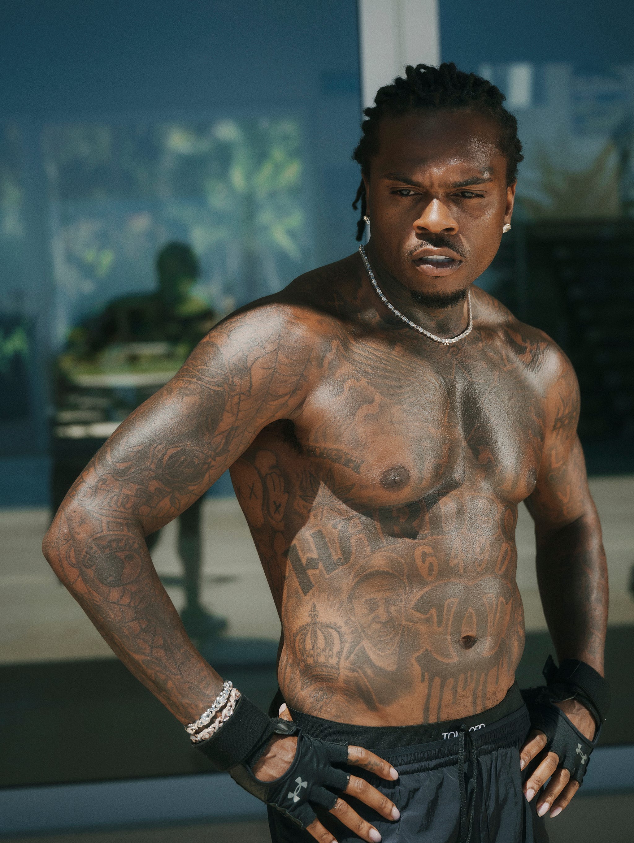 Muscular man with numerous tattoos, standing confidently with a serious expression in front of a glass backdrop.