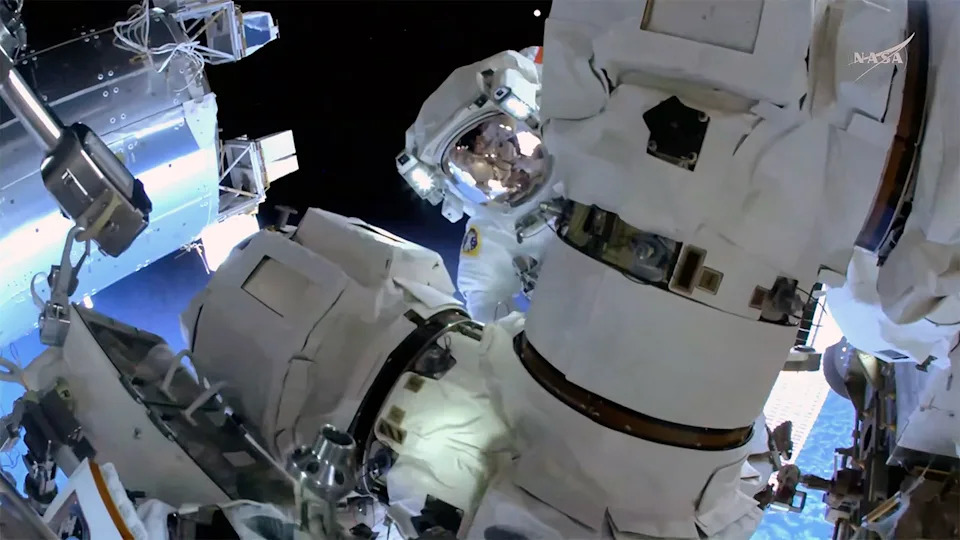 An astronaut in a white spacesuit peers around a robot arm outside of a space station