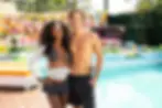 Smiling couple in swimwear pose by a bright resort pool with colorful lounge seating and tropical greenery.