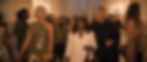 Stylish guests walk through a warm-lit upscale party, led by a blonde woman in gold, a woman in white, and a man in black.