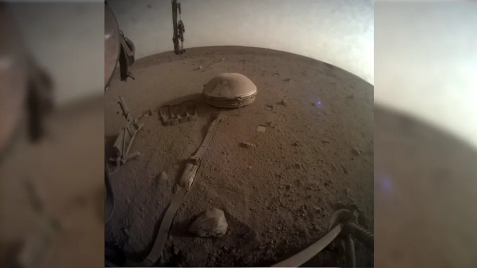 a squat, round lander sits on three metal legs on a reddish-orange dusty surface, with two large wing-like solar arrays, one on each side