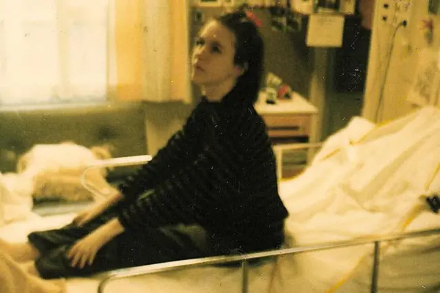 A slightly blurred colour photograph shows Nina sidon for hospital bed for one medical ward. Di bed get raised metal side rails and white bedding, and medical equipment dey mounted on di wall nearby. A large window dey allow daylight from di left side of di image. Cabinets, shelves and hospital furnishings dey seen for di background. Di photograph get di faded colour tones and grainy quality typical of older snapshot from 1989.