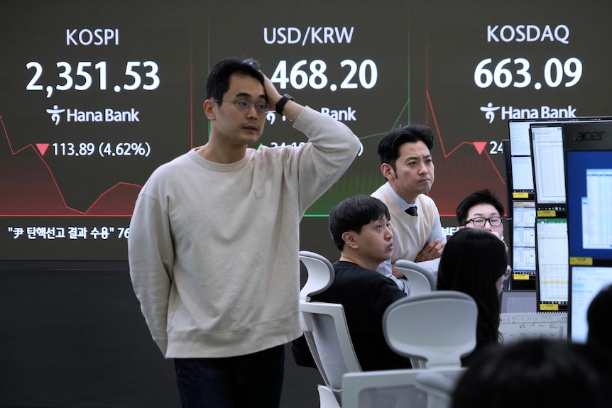 A man holds his hand on his head looking forlorn as numbers on screens behind him flash red showing decline