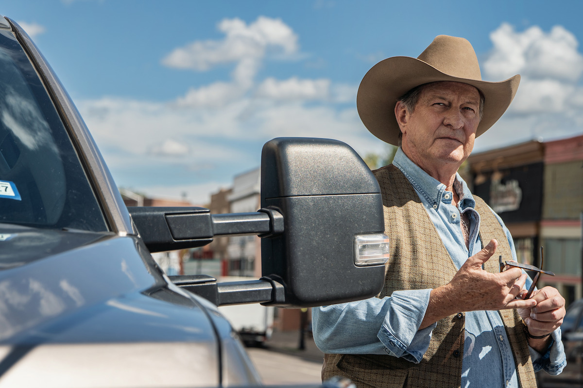 Older man in cowboy hat and vest stands next to pickup truck on a sunny day, small town street with buildings and blue sky in the background, holding sunglasses and looking thoughtful.