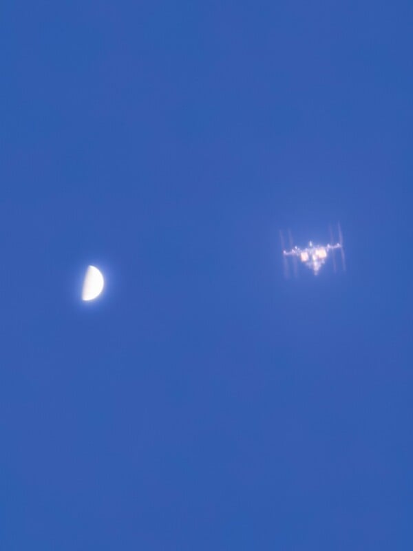 A half moon is visible in a blue sky with the International Space Station appearing nearby, both slightly blurred by atmospheric haze.